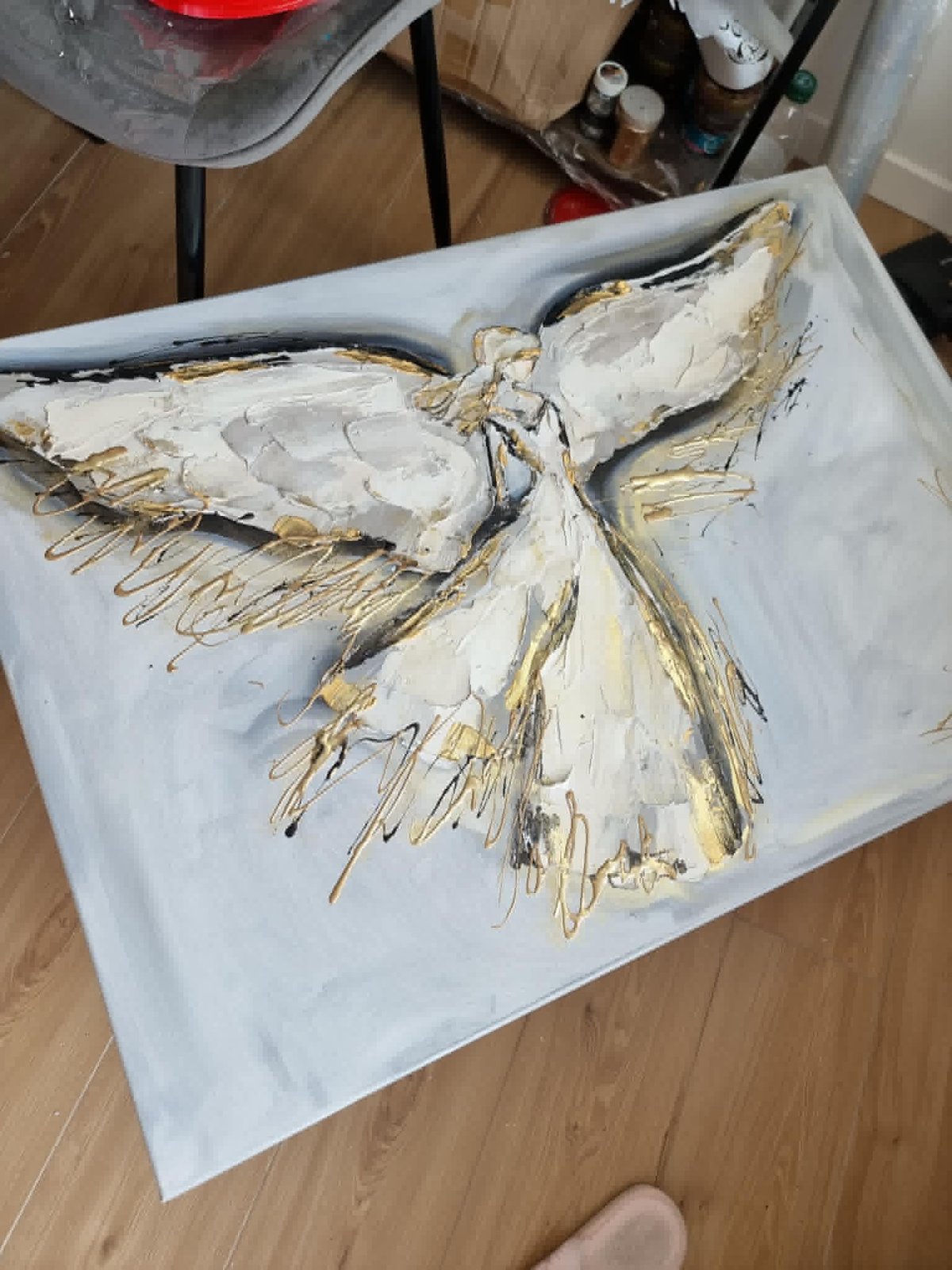 A childs white painted artwork depicting an angel figure with cream and gold colored wings on paper laid on a wooden floor