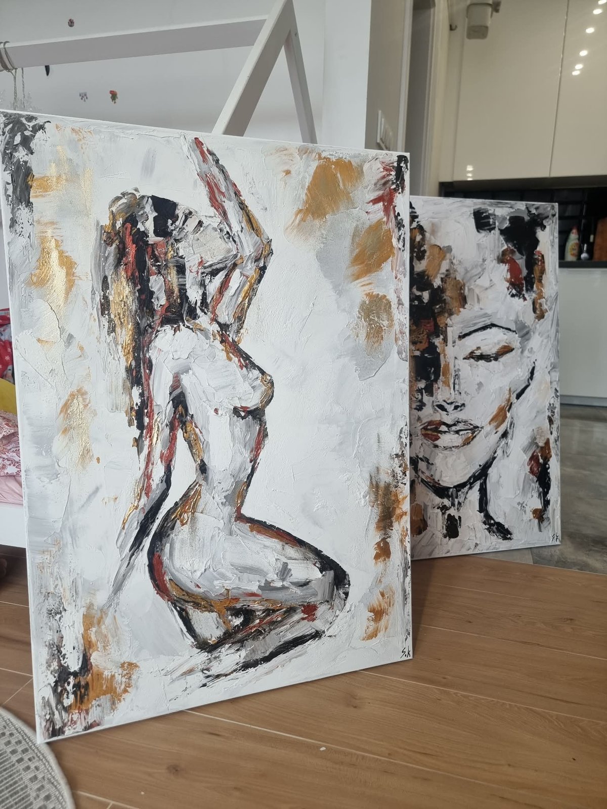 Two abstract painted canvases displayed on wooden floor in gallery space. Left canvas features expressive face profile in earth tones and black. Right canvas shows minimalist portrait.