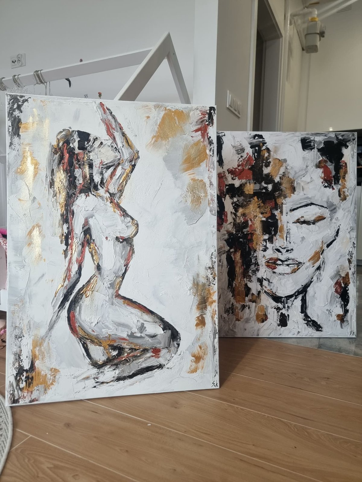 Two abstract mixed media paintings leaning against white wall in artist studio, featuring figure sketches in black, red and gold on canvas