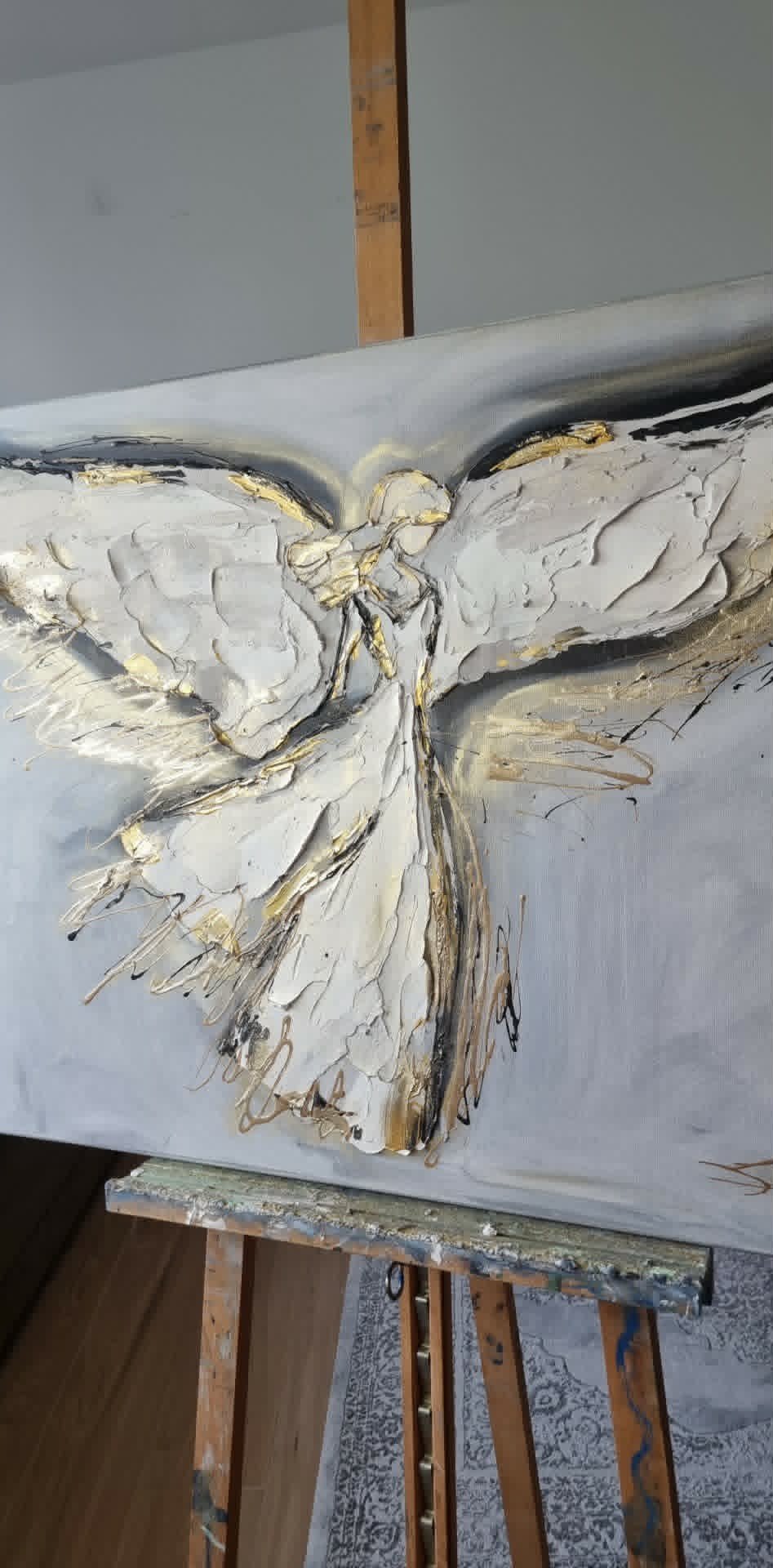 Abstract painting with white, beige, and gold brushstrokes forming a butterfly-like shape on a gray canvas on an easel