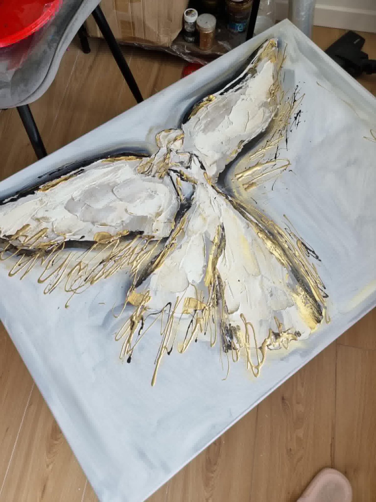 White canvas with a textured painting of an angel or bird figure in white and gold acrylic paint on wooden floor
